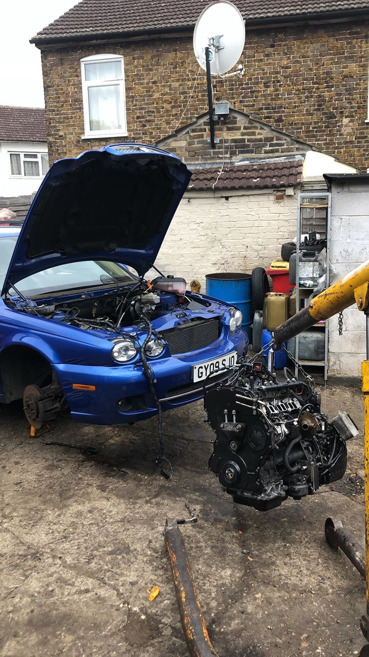 A blue Jaguar X-Type with its bonnet up and the engine lifted out on a crane in the workshop yard