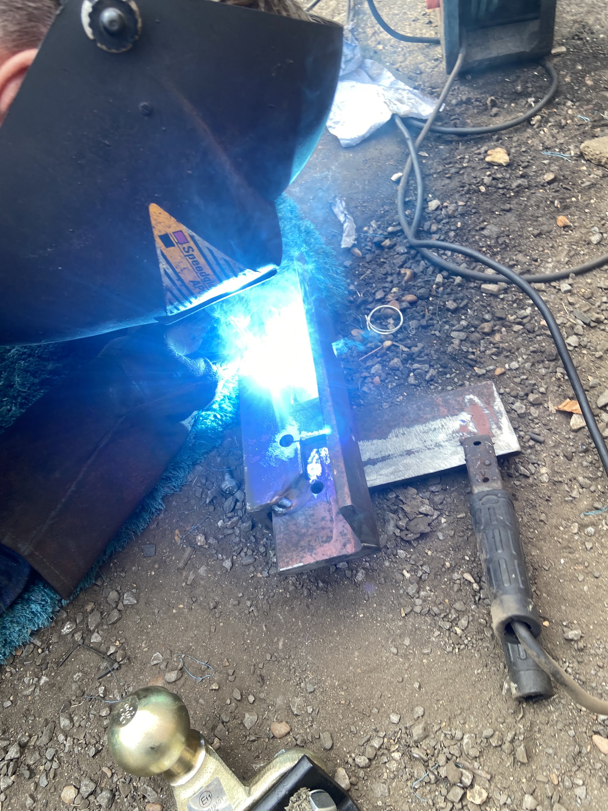 MIG welding a steel bracket on the ground with a tow ball beside it, the welding arc glowing blue